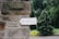 A white arrow-shaped sign with the word 'Wedding' written in elegant, cursive script is attached to a rustic stone wall. The background features lush greenery, including a large tree and various plants.