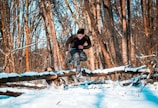 A person is running through a snowy forest, wearing camouflaged pants and a dark long-sleeved shirt. They are leaping over a log that is partially covered in snow. A dog is also present in the scene, energetically engaged in activity. The trees in the background have no leaves, indicating a winter setting.
