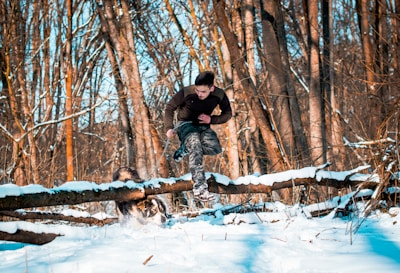 A person is running through a snowy forest, wearing camouflaged pants and a dark long-sleeved shirt. They are leaping over a log that is partially covered in snow. A dog is also present in the scene, energetically engaged in activity. The trees in the background have no leaves, indicating a winter setting.