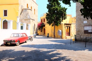 A sunny street scene with vibrant yellow buildings and a classic red car parked on the cobblestone road. There is a motorcycle nearby and lush greenery including trees and bright pink bougainvillea. A small sign reads 'HAMMAM BATHS' on one of the stone walls, hinting at a historical or cultural site.