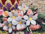 A vibrant and intricate floral arrangement features a mix of natural and crafted elements. Soft pink roses are interspersed with large white woven flowers that have bright yellow centers. Surrounding the arrangement are various other flowers and green foliage, along with textured blooms in red, purple, and cream colors forming a border-like pattern.