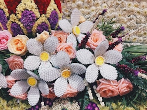 A vibrant and intricate floral arrangement features a mix of natural and crafted elements. Soft pink roses are interspersed with large white woven flowers that have bright yellow centers. Surrounding the arrangement are various other flowers and green foliage, along with textured blooms in red, purple, and cream colors forming a border-like pattern.