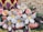 A vibrant and intricate floral arrangement features a mix of natural and crafted elements. Soft pink roses are interspersed with large white woven flowers that have bright yellow centers. Surrounding the arrangement are various other flowers and green foliage, along with textured blooms in red, purple, and cream colors forming a border-like pattern.