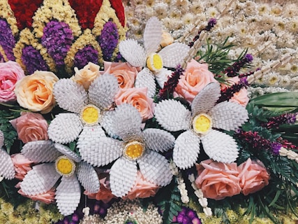 A vibrant and intricate floral arrangement features a mix of natural and crafted elements. Soft pink roses are interspersed with large white woven flowers that have bright yellow centers. Surrounding the arrangement are various other flowers and green foliage, along with textured blooms in red, purple, and cream colors forming a border-like pattern.