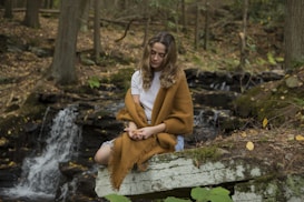 A woman with long hair and wearing a white t-shirt sits peacefully on a stone by a small waterfall in a forest. She is wrapped in a brown blanket and is surrounded by trees and fallen leaves. The scene conveys a sense of tranquility and solitude in nature.