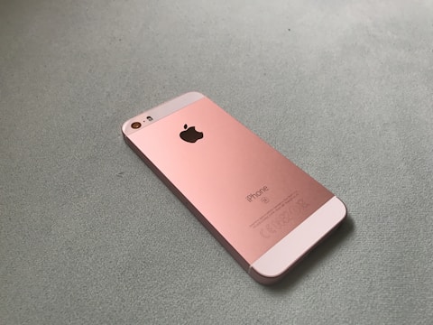 A rose gold smartphone lies on a textured surface. The back of the phone features a prominent Apple logo and text indicating it is an iPhone. The phone's design is sleek and minimalistic.