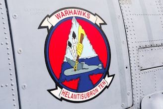 A patch on a metal surface featuring an emblem with the text 'WARHAWKS HELANTISUBRON TEN'. The design includes an iceberg, a submarine, and a bird griping a trident. The main colors are red, blue, white, and black.
