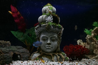 A serene stone sculpture of a meditative face adorned with moss and vines is centered in an aquarium, surrounded by colorful aquatic plants and rocks. A small fish swims nearby, enhancing the tranquil atmosphere.