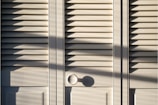 A series of white louvered doors with sunlight casting strong shadows across the surface. The slats create a pattern of light and shadow, and there is a round door handle visible at the bottom right.
