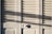 A series of white louvered doors with sunlight casting strong shadows across the surface. The slats create a pattern of light and shadow, and there is a round door handle visible at the bottom right.