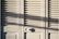 A series of white louvered doors with sunlight casting strong shadows across the surface. The slats create a pattern of light and shadow, and there is a round door handle visible at the bottom right.