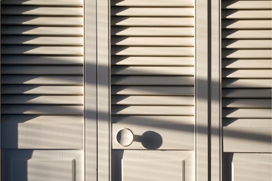 A series of white louvered doors with sunlight casting strong shadows across the surface. The slats create a pattern of light and shadow, and there is a round door handle visible at the bottom right.