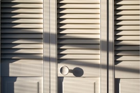 A series of white louvered doors with sunlight casting strong shadows across the surface. The slats create a pattern of light and shadow, and there is a round door handle visible at the bottom right.