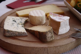 A wooden platter displays an assortment of cheeses, including a wedge of blue cheese, a round of brie, and several other varieties. The cheeses are arranged neatly, with a fork to the side and a plate of salad partially visible in the background.