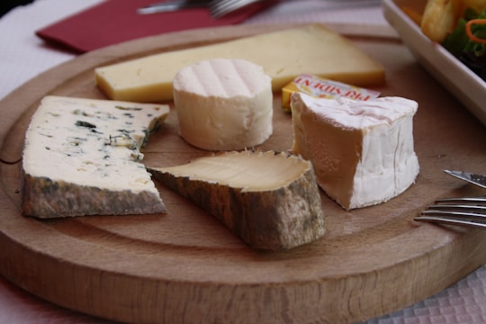 A wooden platter displays an assortment of cheeses, including a wedge of blue cheese, a round of brie, and several other varieties. The cheeses are arranged neatly, with a fork to the side and a plate of salad partially visible in the background.