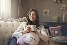A woman with long, wavy hair is sitting on an elegant cream-colored sofa, holding a white mug. She is dressed in a white blouse and dark pants, appearing relaxed and content. Behind her are decorative cushions with geometric patterns and a stack of books on a shelf. Soft lighting adds a cozy atmosphere to the room.
