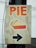 A wooden sign with the word 'PIE' written in large orange letters at the top. Below the text, there is an illustration of a slice of pie showing a golden crust and red filling. At the bottom, a black arrow points to the right.