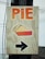 A wooden sign with the word 'PIE' written in large orange letters at the top. Below the text, there is an illustration of a slice of pie showing a golden crust and red filling. At the bottom, a black arrow points to the right.