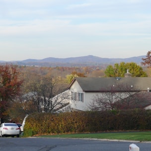 A residential house with a grey roof and white walls, surrounded by trees with autumn leaves and a green hedge. In the background, rolling hills and mountains are visible under a partly cloudy sky. A white car is parked on the left side of the foreground.