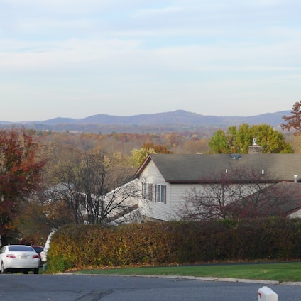 A residential house with a grey roof and white walls, surrounded by trees with autumn leaves and a green hedge. In the background, rolling hills and mountains are visible under a partly cloudy sky. A white car is parked on the left side of the foreground.