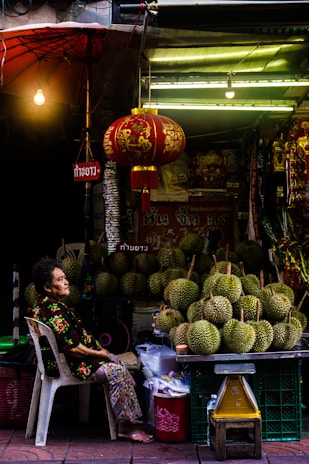 A street vendor is seated beside a stall filled with durians. The stall is decorated with a red lantern and various signs in an Asian script. The vendor, an older woman, is wearing a floral outfit and sits on a white plastic chair. The scene is dimly lit with a warm light bulb hanging from above.