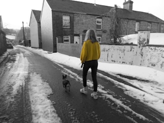 A person wearing a bright yellow jacket walks a small dog on a leash along a snow-covered residential street. The scene is mostly in black and white, highlighting the vibrant color of the jacket against the muted surroundings. Houses with brick exteriors and a slightly overcast sky can be seen in the background, with patches of snow and wet tarmac on the ground.