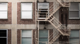 A brick building facade with six rectangular windows. Each window has white blinds, and a metal fire escape with zigzagging stairs runs vertically across the right side. The bricks are a dark red color, with some visible wear and discoloration. Concrete borders separate the windows and add texture to the building's exterior.