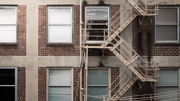 A brick building facade with six rectangular windows. Each window has white blinds, and a metal fire escape with zigzagging stairs runs vertically across the right side. The bricks are a dark red color, with some visible wear and discoloration. Concrete borders separate the windows and add texture to the building's exterior.
