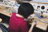 A person wearing a maroon shirt is sitting at a workbench filled with electronic components and tools, assembling or examining small parts. The workspace has organized shelves with various electronic equipment and parts, including wires and a soldering iron.