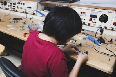 A person wearing a maroon shirt is sitting at a workbench filled with electronic components and tools, assembling or examining small parts. The workspace has organized shelves with various electronic equipment and parts, including wires and a soldering iron.