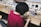 A person wearing a maroon shirt is sitting at a workbench filled with electronic components and tools, assembling or examining small parts. The workspace has organized shelves with various electronic equipment and parts, including wires and a soldering iron.