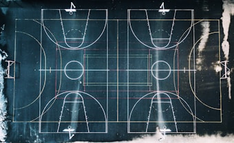 An aerial view of a basketball court with clear demarcations of the playing lines and a slightly worn surface. The lines are white, red, and yellow, contrasting against the dark court background. The court seems to be outdoors, with surrounding areas showing some snow or frost.