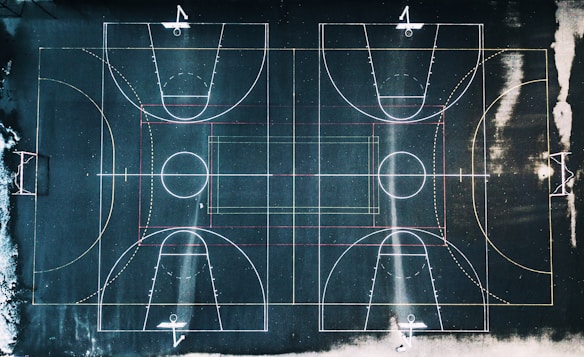 An aerial view of a basketball court with clear demarcations of the playing lines and a slightly worn surface. The lines are white, red, and yellow, contrasting against the dark court background. The court seems to be outdoors, with surrounding areas showing some snow or frost.
