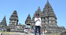A person stands in front of a large stone temple complex with intricate carvings and towering spires. Several other people in casual attire are walking around, some holding umbrellas to shield from the sun. The sky is clear and blue, suggesting a bright and warm day.