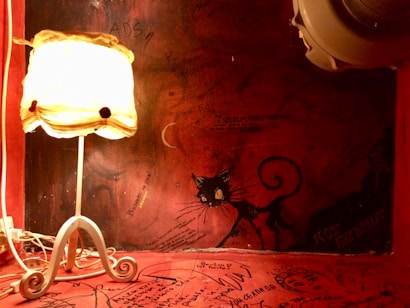 A dimly lit room with a vintage-style lamp on a white ornate stand casting a warm glow. The wall and surface are rich deep red, covered in various handwritten notes and drawings. One prominent drawing on the wall features a black cat with large eyes and an exaggerated, curly tail. The atmosphere is eclectic and artistic.