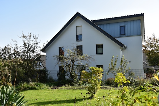 A modern house with white walls and a dark roof is surrounded by a lush garden. The garden features various trees and bushes, with a well-maintained lawn in the foreground. Natural sunlight illuminates the scene, casting gentle shadows alongside.