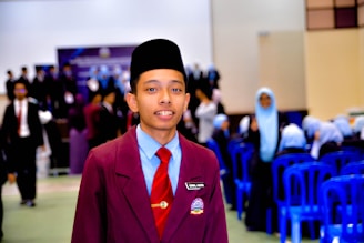 A young person wearing a maroon blazer with a name tag, light blue shirt, red tie, and a traditional black headwear is standing in the foreground. In the background, there are groups of people, some wearing similar attire and others in light blue headscarves, seated on blue plastic chairs. The setting appears to be a formal indoor event, possibly a school or academic gathering.