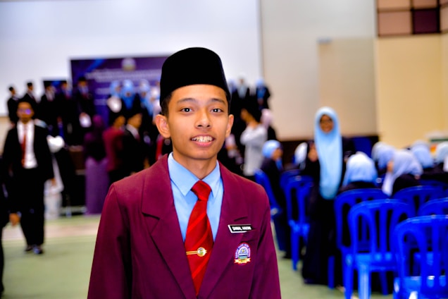 A young person wearing a maroon blazer with a name tag, light blue shirt, red tie, and a traditional black headwear is standing in the foreground. In the background, there are groups of people, some wearing similar attire and others in light blue headscarves, seated on blue plastic chairs. The setting appears to be a formal indoor event, possibly a school or academic gathering.