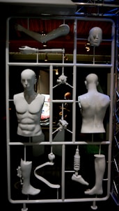 A large, plastic frame contains various disassembled mannequin body parts, including a torso, arms, legs, and a head. The pieces are arranged like a model kit, each connected to the frame. The setting is dimly lit with spotlights highlighting the white figures against a dark background.