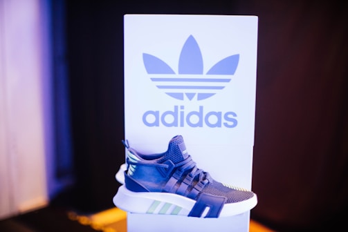 A sneaker displayed in front of a branded backdrop. The shoe has a modern design with a combination of blue, white, and green accents. The background features a large logo and contrasting colors, adding to the focus on the sneaker.