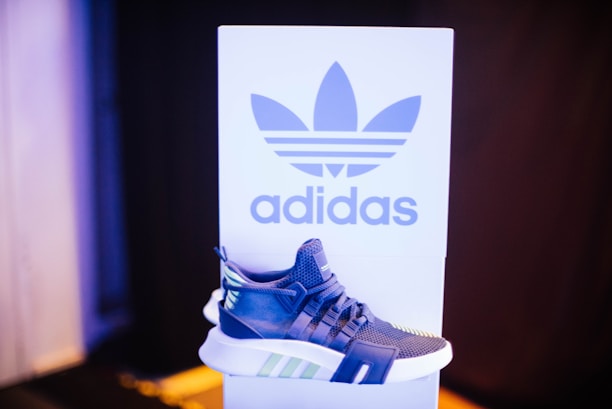 A sneaker displayed in front of a branded backdrop. The shoe has a modern design with a combination of blue, white, and green accents. The background features a large logo and contrasting colors, adding to the focus on the sneaker.