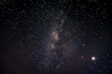 A night sky filled with countless stars, featuring a prominent band of the Milky Way galaxy stretching diagonally across the image. A few bright celestial bodies are visible, with some clouds at the lower left. The vastness and beauty of the universe are showcased against a dark backdrop.