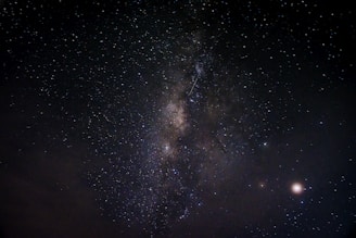 A night sky filled with countless stars, featuring a prominent band of the Milky Way galaxy stretching diagonally across the image. A few bright celestial bodies are visible, with some clouds at the lower left. The vastness and beauty of the universe are showcased against a dark backdrop.