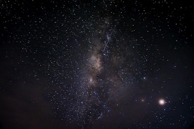 A night sky filled with countless stars, featuring a prominent band of the Milky Way galaxy stretching diagonally across the image. A few bright celestial bodies are visible, with some clouds at the lower left. The vastness and beauty of the universe are showcased against a dark backdrop.