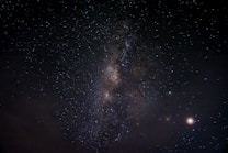 A night sky filled with countless stars, featuring a prominent band of the Milky Way galaxy stretching diagonally across the image. A few bright celestial bodies are visible, with some clouds at the lower left. The vastness and beauty of the universe are showcased against a dark backdrop.