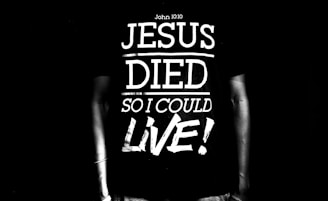A black and white image featuring a person wearing a T-shirt with the text 'Jesus Died So I Could Live!' along with a reference to John 10:10. The text is large and bold, dominating the scene.