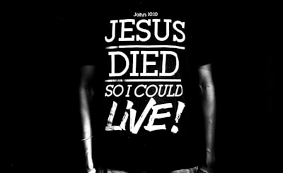 A black and white image featuring a person wearing a T-shirt with the text 'Jesus Died So I Could Live!' along with a reference to John 10:10. The text is large and bold, dominating the scene.