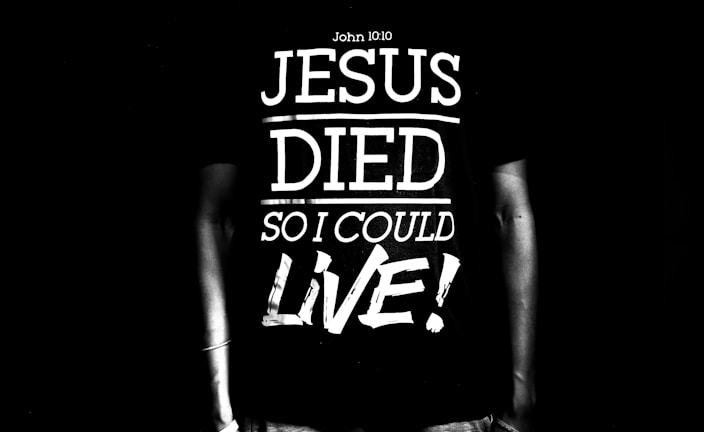 A black and white image featuring a person wearing a T-shirt with the text 'Jesus Died So I Could Live!' along with a reference to John 10:10. The text is large and bold, dominating the scene.