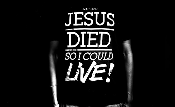 A black and white image featuring a person wearing a T-shirt with the text 'Jesus Died So I Could Live!' along with a reference to John 10:10. The text is large and bold, dominating the scene.
