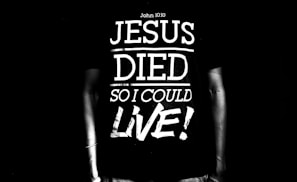 A black and white image featuring a person wearing a T-shirt with the text 'Jesus Died So I Could Live!' along with a reference to John 10:10. The text is large and bold, dominating the scene.